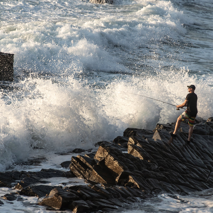 Man standing on the rocky shores of a beach with waves crashing all around him as he fishes